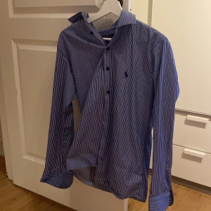 Blåvit randig skjorta från Polo Ralph Lauren - Säljer en klassisk blå och vit randig skjorta från Polo Ralph Lauren med broderad logga på bröstet. Skjortan har långa ärmar och knäppning framtill. Perfekt för dig som gillar stilrena och tidlösa plagg. Det finns en defekt på den, en knapp har lossnat på ärmen, som ni ser på sista bilden!