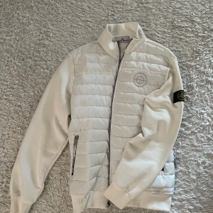 Vit cardigan från Stone Island - Säljer nu en vit cardigan från Stone Island i väldigt bra skick, knappt använd med broderad logga på bröstet och klassisk patch på ärmen. Jackan har dragkedja framtill, ståkrage och quiltade paneler på framsidan. Perfekt för kyliga dagar.