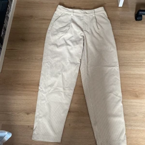 Beiga manchesterbyxor från KappAhl - Snygga beiga manchesterbyxor från KappAhl med klassisk rak passform och hög midja. Byxorna har diskreta fickor fram och stängs med knapp och dragkedja. Perfekta för en stilren och avslappnad look.