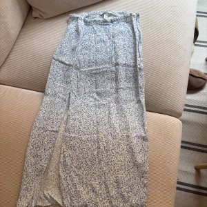 Blåvit blommig långkjol - Säljer en långkjol från H&M i vitt tyg med blått blommigt mönster. Kjolen har rak passform med slits.