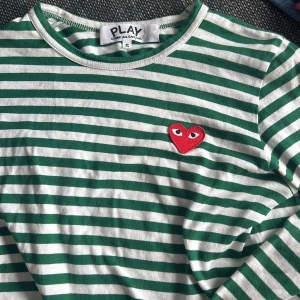 Randig långärmad tröja från Comme des Garçons Play - Grön- och vitrandig långärmad tröja från Comme des Garçons Play med det ikoniska röda hjärtat broderat på bröstet. Klassisk rund halsringning och mjukt bomullsmaterial. Perfekt för en stilren och trendig look.