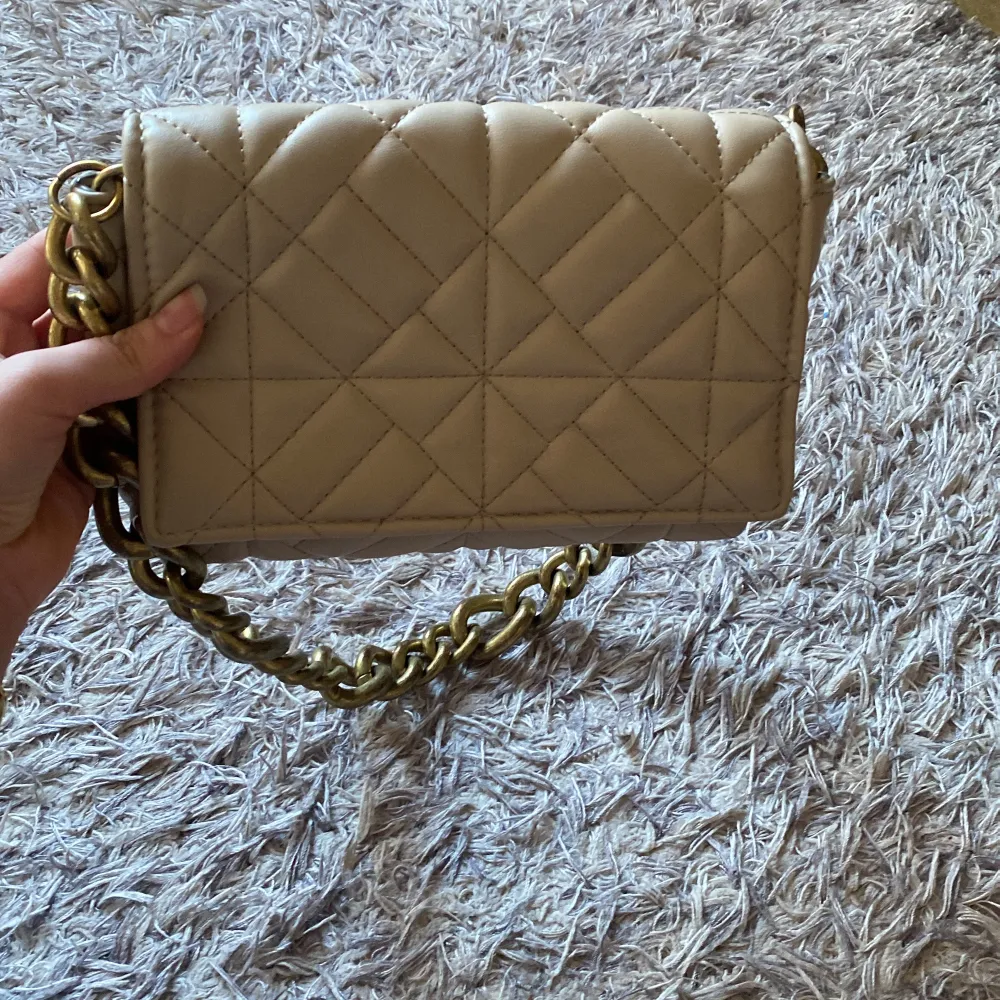 Snygg beige axelväska med quiltat mönster och en rejäl guldfärgad kedja som axelrem. Väskan har ett praktiskt magnetlås och ett rymligt inre fack. Perfekt för att ge din outfit en stilfull touch. Kan gå ner i pris vid snabb affär💖. Laukut & Käsilaukut.
