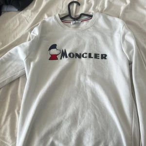 Vit långärmad tröja från Moncler - Säljer en stilren vit långärmad tröja från Moncler med deras ikoniska logotyp tryckt på framsidan. Perfekt för en avslappnad och trendig look. Tröjan har en rund halsringning och är tillverkad i ett mjukt material.
