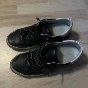 Svarta sneakers från Philippe Model - Säljer ett par svarta sneakers från Philippe Model med vita detaljer på hälen och svarta snören med text. Skorna har en klassisk rund tå och är tillverkade i skinn. Perfekta för dig som gillar stilrena och bekväma skor.