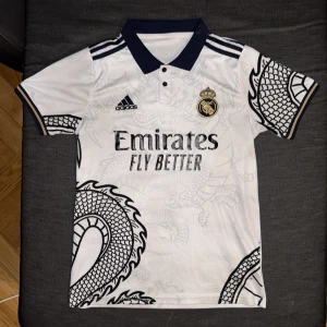 Real Madrid fotbollströja med drakmönster - Vit Real Madrid fotbollströja från Adidas med svart krage och svarta detaljer. Tröjan har ett stort drakmönster i ljusgrått över hela framsidan och baksidan samt klubbmärke och Adidas-logga på bröstet. Sponsortexten 'Emirates Fly Better' syns tydligt.