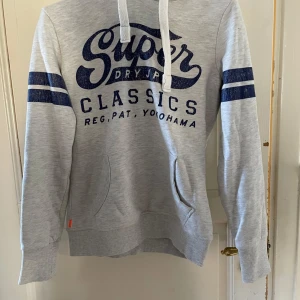 Grå hoodie från Superdry - Säljer en grå hoodie från Superdry med blå text och ränder på ärmarna. Tröjan har en känguruficka och vita dragsnören vid huvan. Perfekt för en avslappnad stil.