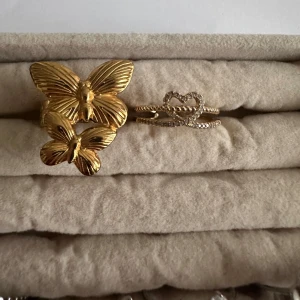 Guldfärgade fjärilsringar - Två vackra guldfärgade ringar med fjärilsdesign. Den ena ringen har två fjärilar i olika storlekar, medan den andra har en elegant hjärtformad detalj med glittriga stenar. Perfekta för att ge en touch av elegans till din stil.