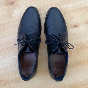 Svarta skinnskor med snörning - Klassiska svarta skor i skinn  med rund tå och snörning. Skorna har en elegant och stilren design med bruna innersulor och låg klack. Perfekta för en dressad look. Nyskick, endast använda två gånger.