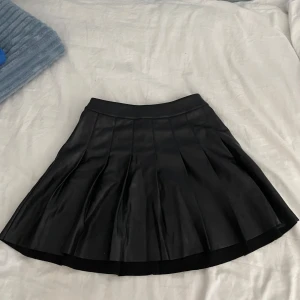 Svart kortkjol från ONLY - Snygg svart kortkjol från ONLY i ett läder/skinn material. Kjolen har en dragkedja på baksidan och veckad design som ger en stilren look. Aldrig använd. Perfekt för att skapa en chic outfit!