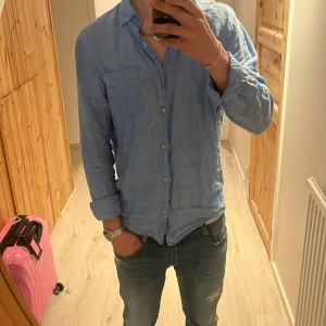 Blå linneskjorta från Kappahl - Snygg blå linneskjorta från Kappahl med lång ärm och klassisk krage. Skjortan har knappar framtill och en bröstficka. Perfekt för en avslappnad stil.