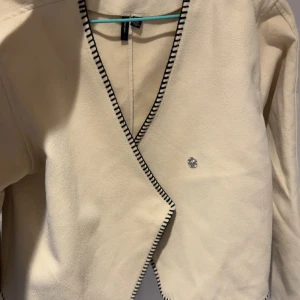 Beige omlottjacka från Vero Moda - Säljer en beige omlottjacka från Vero Moda med svartvitt kantband och en dekorativ knapp framtill. Jackan har lång ärm och en stilren, minimalistisk design som passar perfekt till vår och höst.