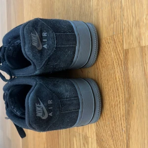 Svarta Nike Air Force 1 sneakers - Säljer ett par svarta Nike Air Force 1 sneakers med svart sula och klassisk Nike-logga på sidan. Skorna har snörning och är tillverkade i mocka med en stilren och tidlös design. Perfekta för dig som gillar streetwear och vill ha en ikonisk sneaker. Dem är lite slitna men funkar perfekt och super skörna 