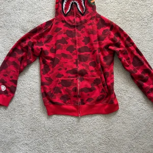 Röd camouflage zip hoodie från BAPE - Säljer en röd BAPE  hoodie med dragkedja och hajmotiv på huvan. Huva med broderade detaljer och bokstäverna WGM samt tigerpatch. Perfekt statement-plagg för streetwear-stilen.