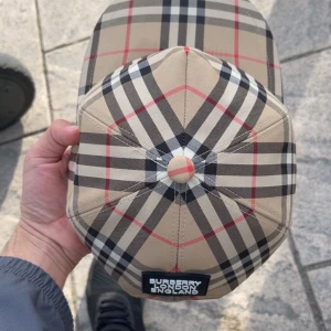 Beige rutig keps från Burberry - Snygg keps från Burberry i klassiskt beige rutigt mönster med svart, vitt och rött. Justerbar baksida med svart spänne och broderad Burberry London England-logga. Perfekt accessoar för en trendig look. Äkta burberry