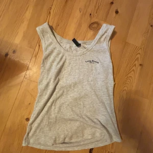 Ljusgrå ribbad linnetopp från Divided - Säljer en ljusgrå ribbad linnetopp från Divided med liten text 'Long Beach' på bröstet. Toppen är ärmlös och har en rund halsringning. Perfekt för varma dagar eller som lager under andra plagg.