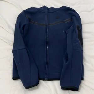 Blå zip-hoodie från Nike - Mörkblå hoodie från Nike med dragkedja framtill och huva. Jackan har svarta detaljer längs bröstet och på ena ärmen, samt fickor med dragkedja. Perfekt för träning eller chill dagar.
