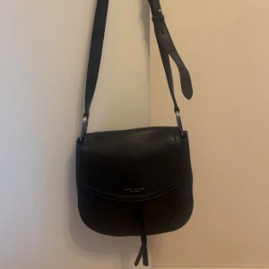 Svart axelväska från Marc Jacobs - Snygg svart axelväska från Marc Jacobs i skinn med justerbart axelband och lock med diskret logga framtill. Väskan har en rundad form och innerfack med dragkedja. Perfekt för att bära det viktigaste med stil.