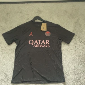 PSG fotbollströja  - Säljer en svart PSG fotbollströja med rosa detaljer och Qatar Airways som sponsor. Tröjan har korta ärmar, Paris Saint-Germain klubbmärke på bröstet och Jordan-logga. Materialet är lätt och andas, perfekt för fotboll eller träning.