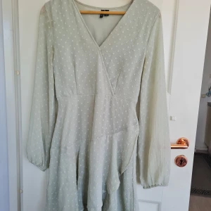 Ljusgrön omlottklänning med prickigt mönster från Vero Moda - Supersöt ljusgrön omlottklänning från Vero Moda med diskret prickigt mönster. Klänningen har lång ärm, v-ringning och knytband i midjan för en fin passform. Perfekt för vår och sommar!