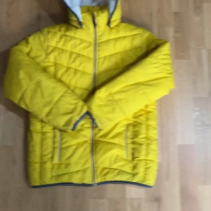 Gul pufferjacka med huva från Name It - Snygg gul pufferjacka från Name It i storlek 152 (12 år). Jackan har huva, dragkedja framtill och två fickor med dragkedja. Perfekt för kyligare dagar och har en sportig look med randiga detaljer vid ärmslut och nederkant.