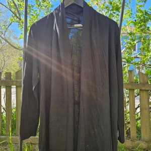 Svart lång cardigan kofta - Svart lång cardigan kofta med öppen front och långa ärmar. Perfekt för lager-på-lager och enkel att matcha med olika outfits. Tunnare material som passar bra till mildare väder.