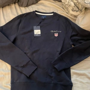 Mörkblå sweatshirt från GANT - Säljer en mörkblå sweatshirt från GANT med rund halsringning och broderad logotyp samt sköld på bröstet. Tröjan har långa ärmar och ribbade muddar vid ärmslut och nederkant. Perfekt för en stilren och avslappnad look.oavnädn nypris 999