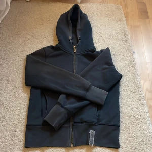 Svart hoodiejacka från Sail Racing - Svart hoodiejacka från Sail Racing med dragkedja framtill, två fickor och justerbar huva. Tjock tröjan har ribbade muddar och en liten Sail Racing-detalj nertill. Perfekt för en sportig och avslappnad stil.