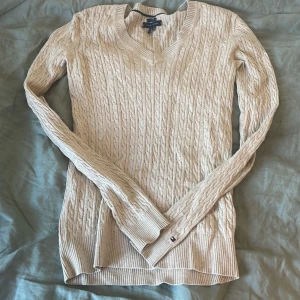 Beige kabelstickad tröja från Tommy Hilfiger - Säljer en beige kabelstickad tröja från Tommy Hilfiger i pima cotton. Tröjan har v-ringning, långa ärmar och en liten broderad logga vid ärmslutet. Perfekt för lager-på-lager och snygg till jeans eller kjol.