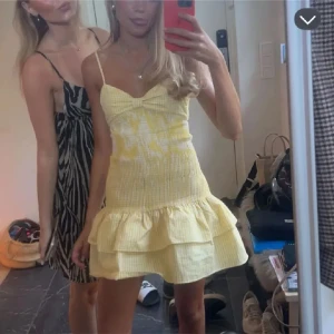 Gul klänning - Superfin gul kortklänning med rutigt mönster och smockad överdel. Klänningen har tunna axelband, volangdetaljer nertill och en söt rosett framtill. Perfekt för varma dagar och somriga tillfällen. 