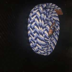 Flätat blått och beige bälte med bruna skinn-detaljer - Säljer ett snyggt flätat bälte i blått och beige med bruna skinn-detaljer vid spännet och änden. Bältet har ett klassiskt metallspänne och passar perfekt till jeans eller chinos.