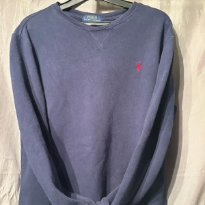 Mörkblå sweatshirt från Polo Ralph Lauren - Mörkblå sweatshirt från Polo Ralph Lauren med klassisk rund halsringning och röd broderad logga på bröstet. Tröjan har långa ärmar och är tillverkad i mjukt bomullsmaterial. Perfekt för en avslappnad och stilren look. Skulle påstå att denna tröja passar någon som har S i storlek. 