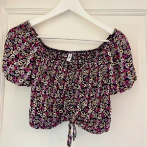 Blommig croppad blus med puffärmar - Superfin croppad blus med blommigt mönster i rosa, vit och gul på svart botten. Puffiga kortärmar och knytning framtill. I mycket bra skick!