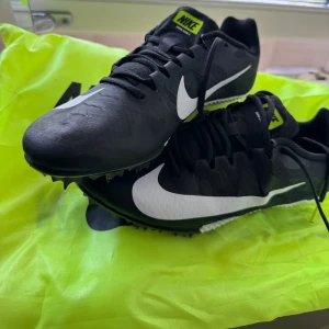 Svarta Nike löparskor med spik (Zoom Rival S) - Säljer ett par svarta Nike löparskor med vita detaljer och spikad sula. Skorna har snörning, tydlig Nike-logga och texten 'Nike Racing' bak. Perfekta för löpning på bana. Kommer med originalpåse. 