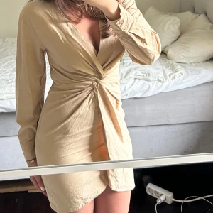 Beige omlottklänning med lång ärm - Snygg beige omlottklänning med lång ärm och v-ringning. Klänningen har en twist-detalj framtill som ger en smickrande passform och en kort längd. Perfekt för dig som gillar stilrena och trendiga plagg.