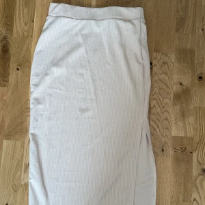 Beige ribbad långkjol - Säljer en beige knälångkjol med diskret ribbad struktur. Kjolen har en slits på sidan som ger en snygg detalj och är perfekt för en stilren look. Storlek s