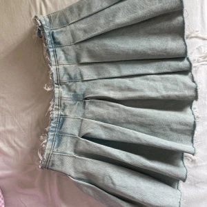 Ljusblå plisserad jeanskjol - Säljer en ljusblå plisserad jeanskjol med råa kanter upptill. Kjolen har en cool och avslappnad vibe med tydliga sömmar och breda veck. Perfekt för dig som gillar streetstyle och denim.
