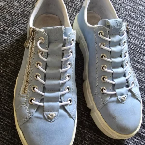 Ljusblå sneakers med dragkedja  - Säljer ett par ljusblå sneakers med vita snören och dragkedja på sidan. Skorna har rund tå, platt sula och är tillverkade i mocka med vita detaljer.