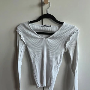 Vit ribbad långärmad topp från NA-KD - Säljer en vit ribbad långärmad topp från NA-KD i storlek XS. Toppen har v-ringning och små volangdetaljer vid axlarna. Perfekt att styla till olika outfits och passar året runt.
