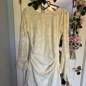 Vit/cremefärgad långärmad klänning med blommigt mönster från Boohoo - Elegant vit/cremefärgad klänning från Boohoo med lång ärm och subtilt blommigt mönster i tyget. Klänningen har en v-ringad öppen rygg och lätt rynkade detaljer framtill. Perfekt för speciella tillfällen och ger en romantisk känsla.