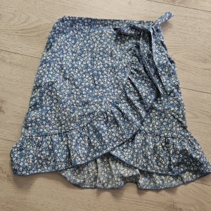 Blå blommig omlottkjol med volang - Supersöt blå omlottkjol med vitt blommönster och volangkant. Kjolen knyts i midjan med en rosett och har dragkedja. Litet hål i en sömm som inte syns alls när den är på. 💞Storlek xs