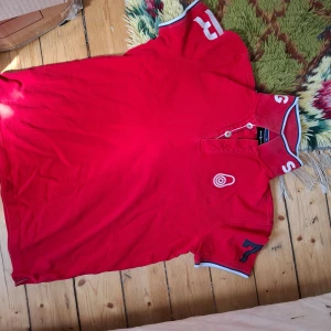 Röd piké t-shirt med tryck och detaljer - Röd piké t-shirt från Paul & Shark med vita och blå detaljer på krage och ärmslut. Snyggt tryck med siffror och text på kragen samt ärmen. Klassisk pikékrage med knappar och broderad logga på bröstet.