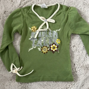 Grön långärmad topp med knyt och blommor - Snygg grön långärmad topp med vita knytband vid hals och ärmslut. Tryck framtill med text och blommor i gult och brunt. Är egentligen barnkläder men vem gillar inte vintage y2k toppar?? Skriv vid frågor och funderingar🤩🤩✊✊🤯🤯