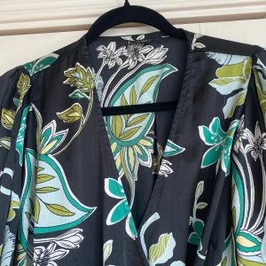 Blommig långklänning från Gina Tricot - Säljer en svart långklänning från Gina Tricot med stora blommiga mönster i grönt, turkost och vitt. Klänningen har v-ringning, trekvartsärmar med lätt puff och markerad midja. Perfekt för dig som gillar färgstarka prints och flowy stil.