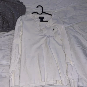 Vit långärmad tröja från Polo Ralph Lauren - Vit långärmad tröja från Polo Ralph Lauren med klassisk krage och knappar vid halsen. Tröjan har det ikoniska broderade logotypen på bröstet och är tillverkad i mjuk bomull. Perfekt för en stilren och avslappnad look.