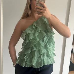 Grön volangig oneshoulder-top - Säljer en ljusgrön oneshoulder-top med flera lager volanger. Toppen har en asymmetrisk axel och dragkedja på sidan. Perfekt för dig som vill sticka ut med en unik och trendig look💚