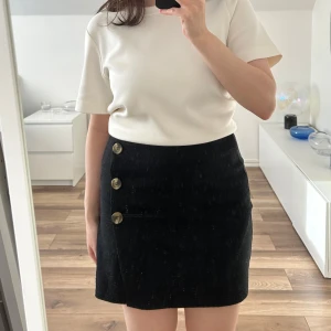 Svart kjol med knappar - Säljer en svart kjol med tre stora dekorativa knappar framtill. Kjolen har en rak passform.