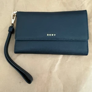 DKNY plånbok - Snygg svart plånbok från DKNY i stilren design med guldfärgade detaljer och avtagbar handledsrem. Plånboken har flera kortfack, dragkedjefack på baksidan och tryckknappsstängning. Perfekt för att hålla ordning på kort och kontanter.