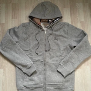 Grå hoodie från Burberry med dragkedja - Säljer en grå hoodie från Burberry med dragkedja och klassiskt rutigt foder i huvan. Tröjan har långa ärmar, två fickor framtill och justerbar huva med snören. Perfekt för en stilren och bekväm look.