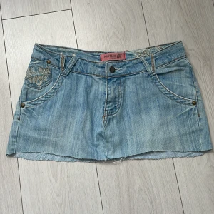 Ljusblå jeanskjol med broderade fickor - Säljer en ljusblå jeanskjol från YMI Jeanswear med snygga broderier i beige på både fram- och bakfickor. Kjolen har klassisk femficksdesign, rå kant nertill och knappstängning framtill. Perfekt för dig som gillar detaljer och denim.