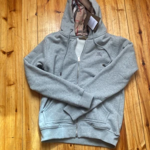 Grå hoodie med dragkedja från Burberry - Säljer en stilren grå hoodie från Burberry med dragkedja och klassiskt rutigt foder i huvan. Tröjan har fickor framtill och justerbar huva med snören. Perfekt för en avslappnad och snygg look.
